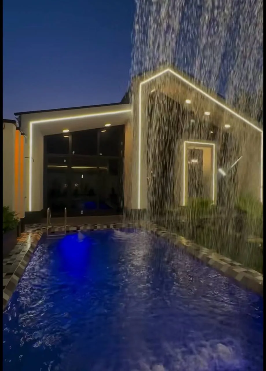 Pool and pavilion shell under construction, then night-lit finished pool with waterfall and LED roof edge.