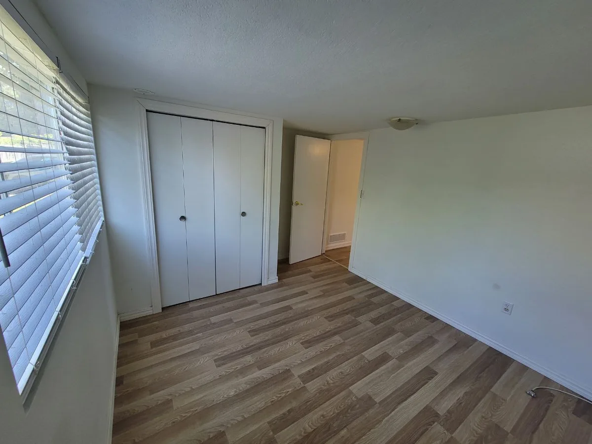 Lower drywall cut back exposing insulation on the same light plank floor, then finished bedroom with closet and blinds.