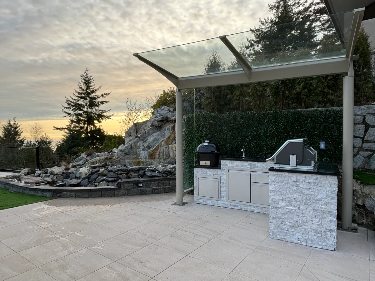 Patio hardscape with kitchen structure materials staged on site, then completed stone-clad kitchen under the canopy.