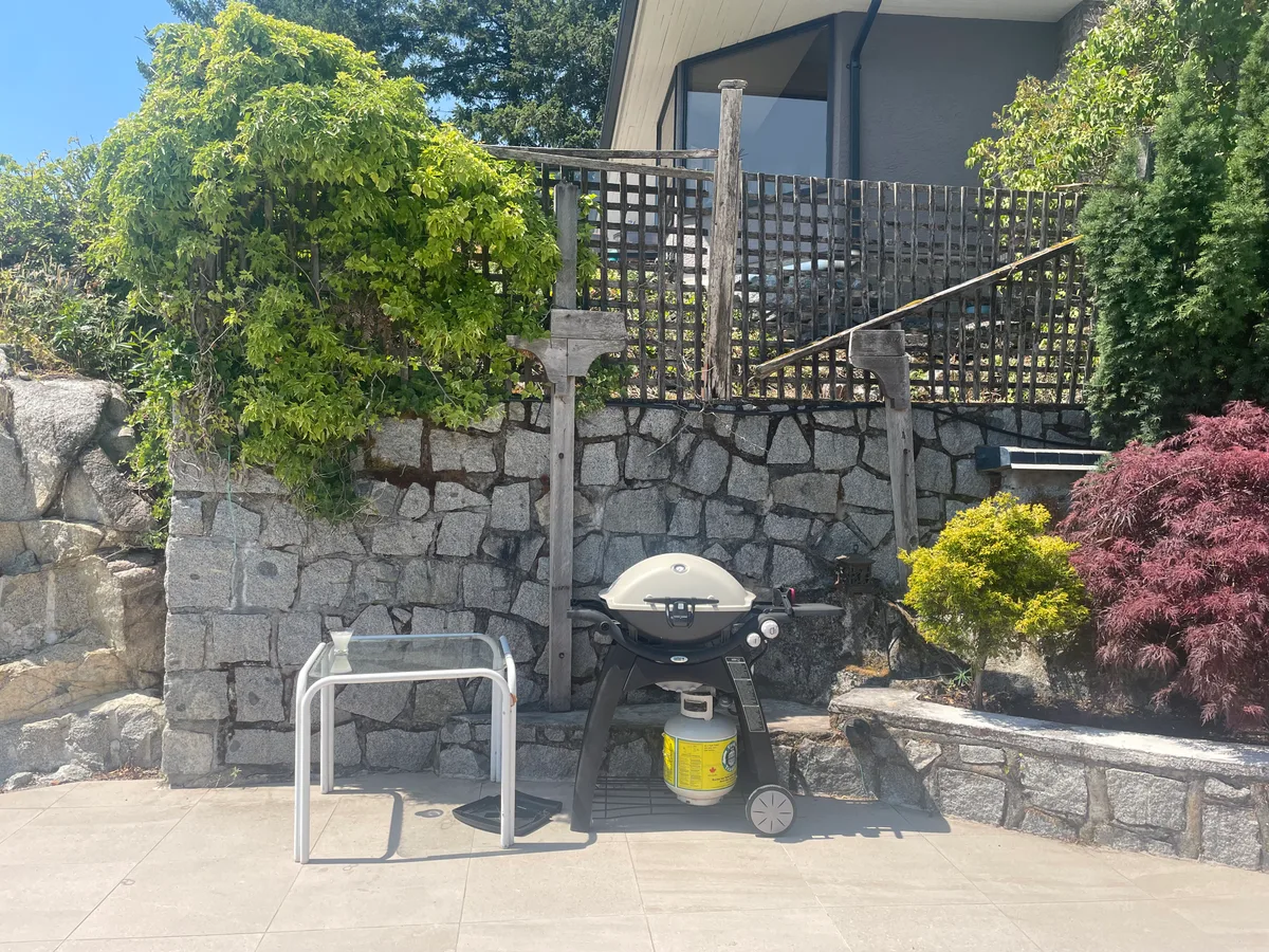 West Vancouver Outdoor Kitchen & Glass Canopy — before — Same stone-wall cooking corner: portable grill and side table, then built-in outdoor kitchen with stainless appliances and stone veneer.