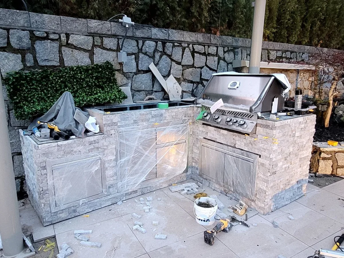 West Vancouver Outdoor Kitchen & Glass Canopy — before — Outdoor kitchen island mid-install with protective film on cabinets, then finished counters, sink, and unwrapped stainless fronts.