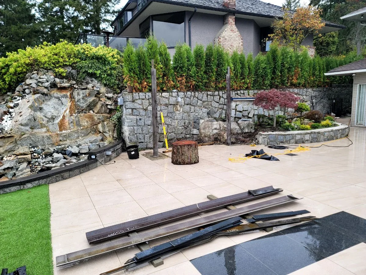 West Vancouver Outdoor Kitchen & Glass Canopy — before — Patio hardscape with kitchen structure materials staged on site, then completed stone-clad kitchen under the canopy.