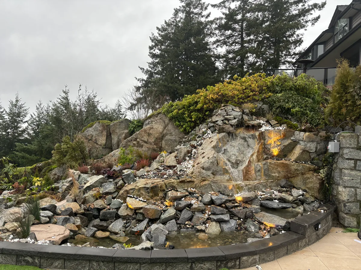 Raw stacked rock face during construction, then completed hillside waterfall with surrounding plantings.