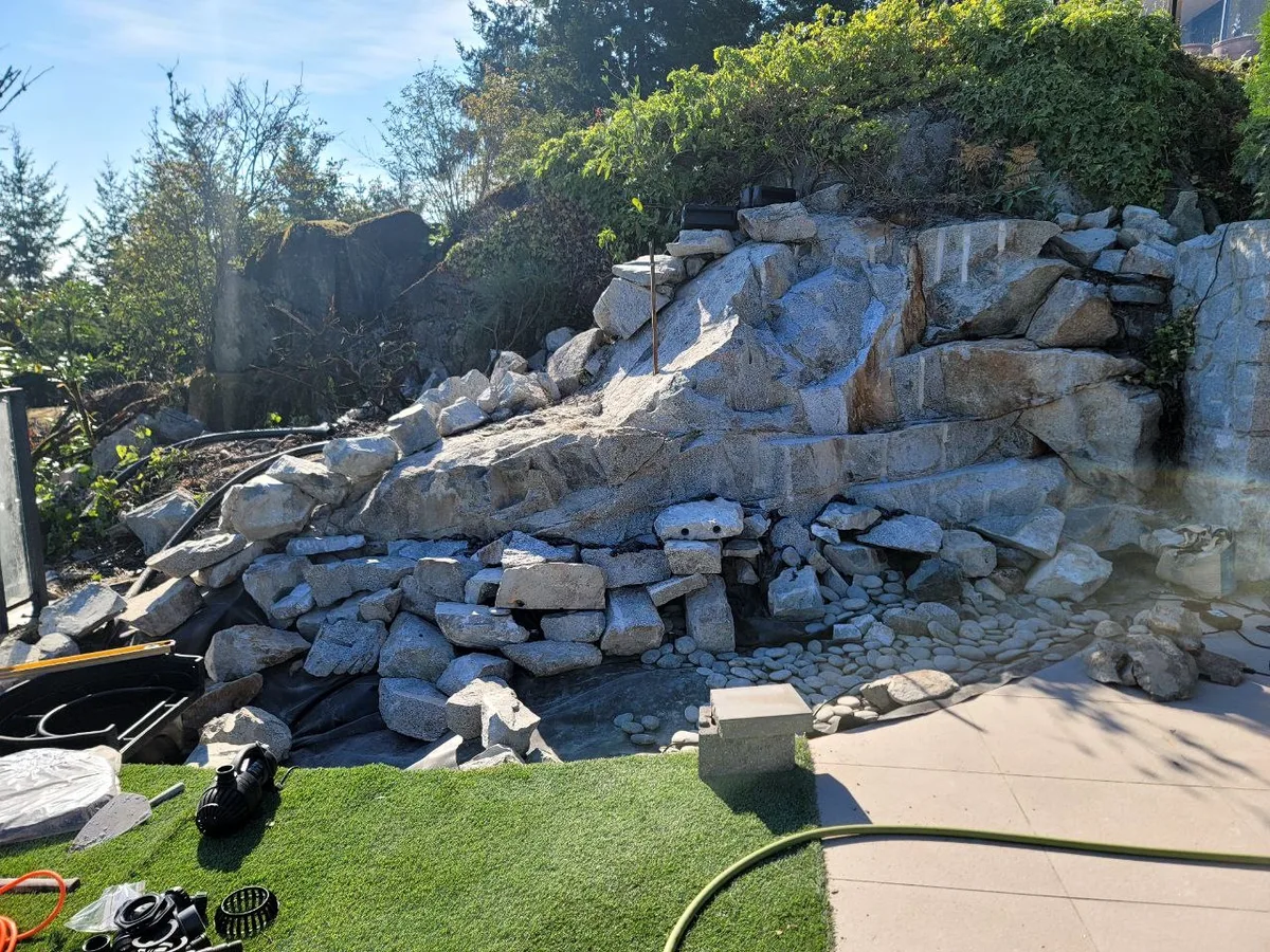 West Vancouver Rock Waterfall & Hillside Hardscape — before — Waterfall rockwork with liner, pump parts, and tools exposed, then finished cascading feature with integrated landscape lighting.