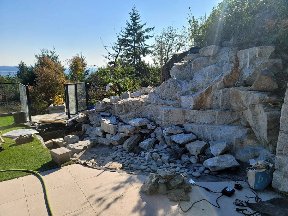 West Vancouver Rock Waterfall & Hillside Hardscape — before — Waterfall under construction beside glass railing and ocean-view trees, then finished flowing rock cascade.